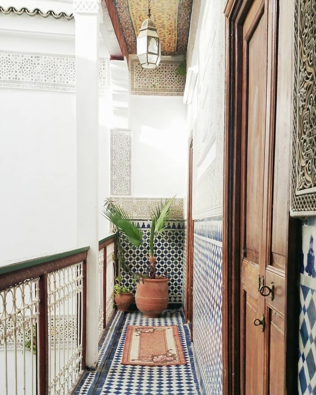Auto-generated description: A beautifully tiled corridor features decorative patterns, ornate woodwork, potted plants, and a traditional lantern hanging from the ceiling.