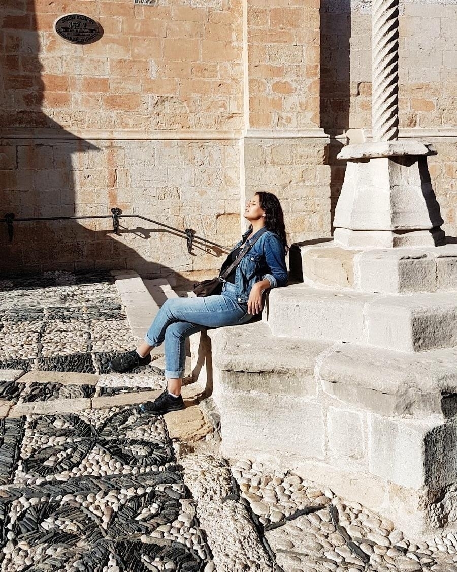 Auto-generated description: A person sits on stone steps, basking in the sunlight against a backdrop of textured stone architecture.
