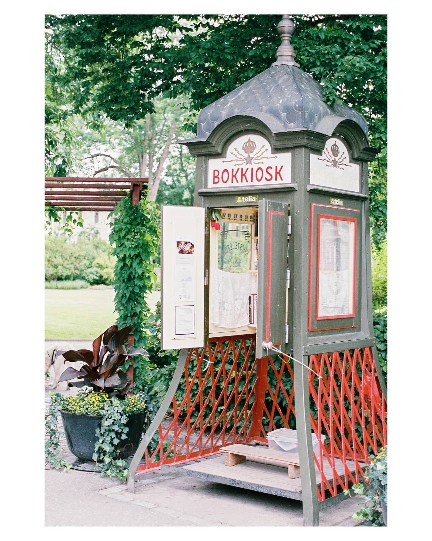 Auto-generated description: A charming old-fashioned kiosk, labeled BOKKIOSK, is surrounded by greenery and plants.