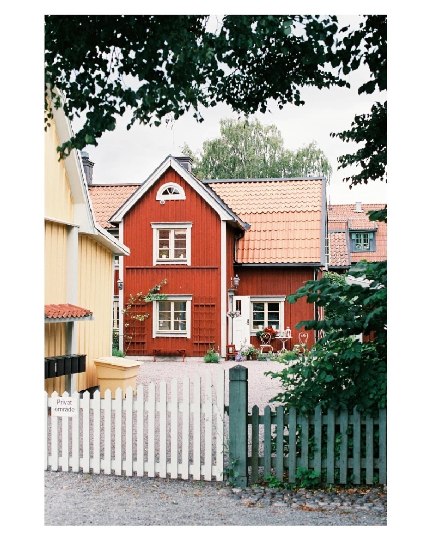 Auto-generated description: A charming red wooden house with a tiled roof sits behind a white picket fence and is surrounded by greenery.