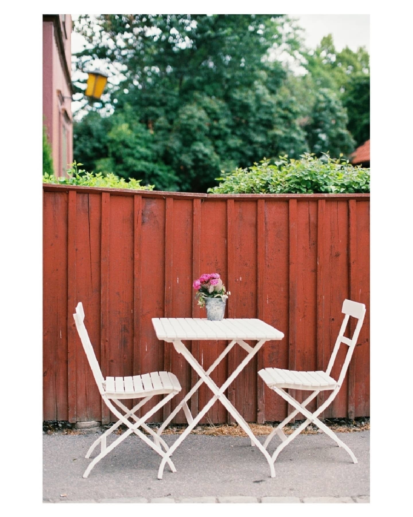 Auto-generated description: A small outdoor table with two white chairs and a vase of flowers is set against a rustic red wooden fence.