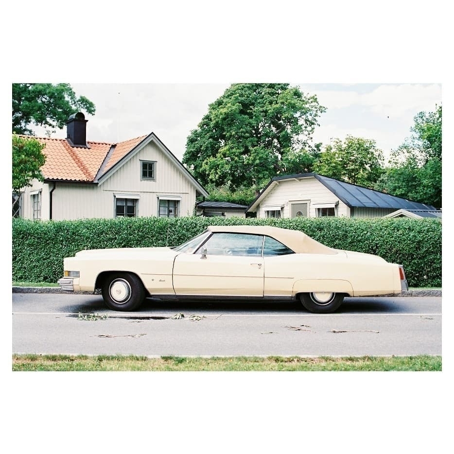 Auto-generated description: A vintage yellow convertible car is parked on a street in front of two quaint houses with lush greenery in the background.