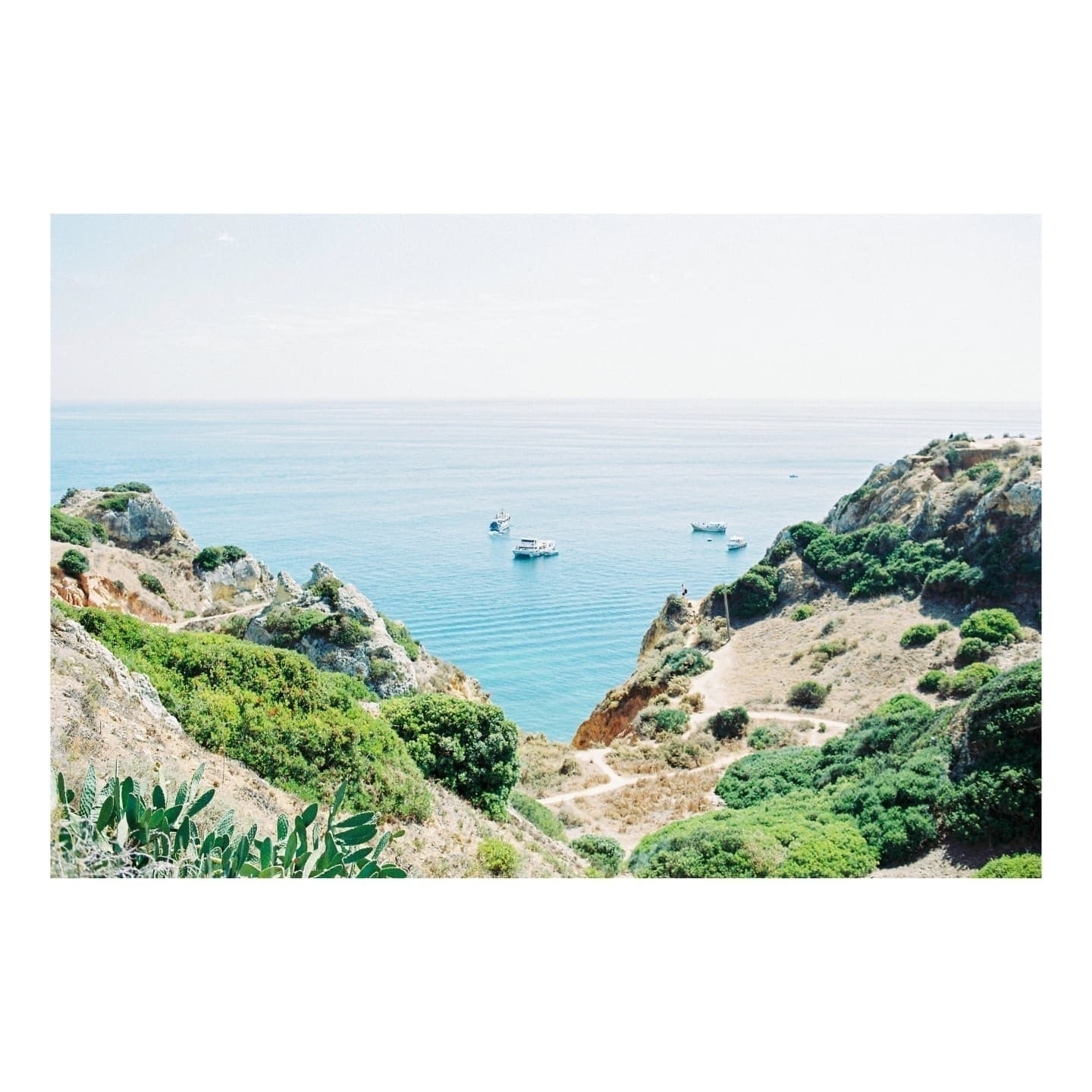 Auto-generated description: A picturesque coastal scene features rugged cliffs and lush greenery overlooking crystal-clear blue waters with a few boats gently floating.