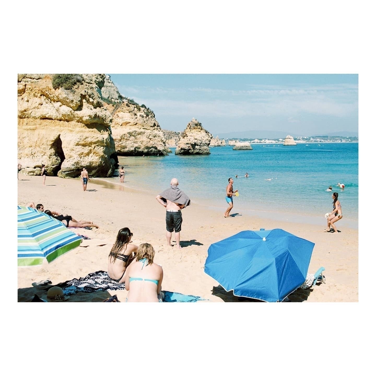 Auto-generated description: People enjoy a sunny day at a sandy beach with rock formations and blue umbrellas.