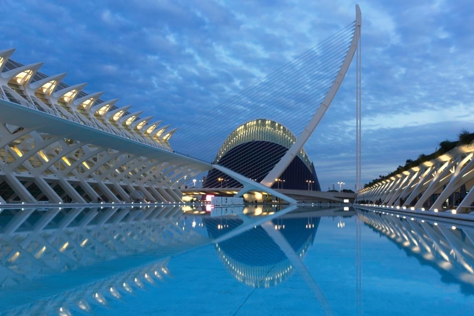 Auto-generated description: A futuristic architectural complex with distinctive curved structures and a large reflecting pool is set against a cloudy evening sky.