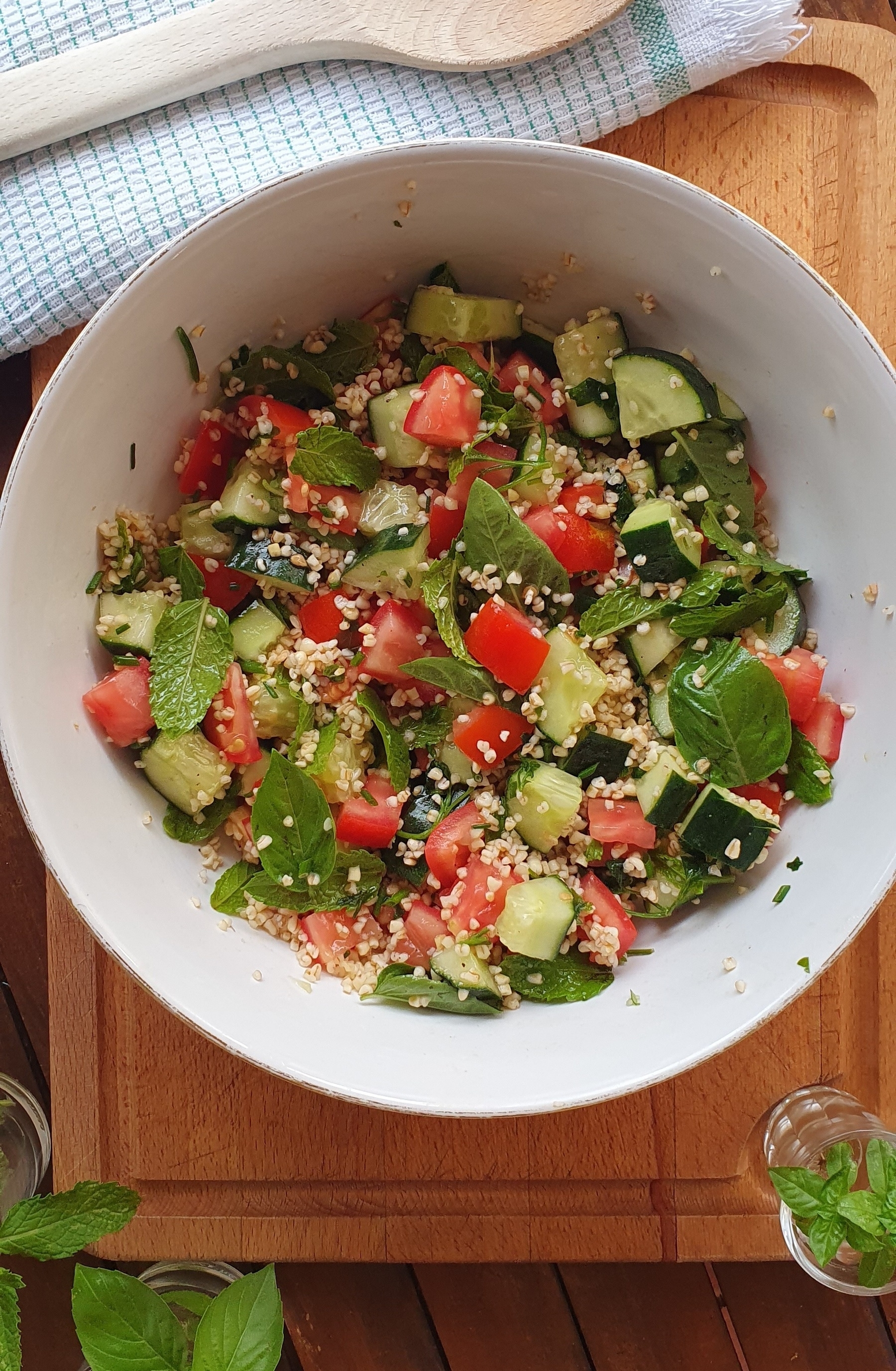 Auto-generated description: A fresh salad with tomatoes, cucumbers, mint, and grains is served in a white bowl.