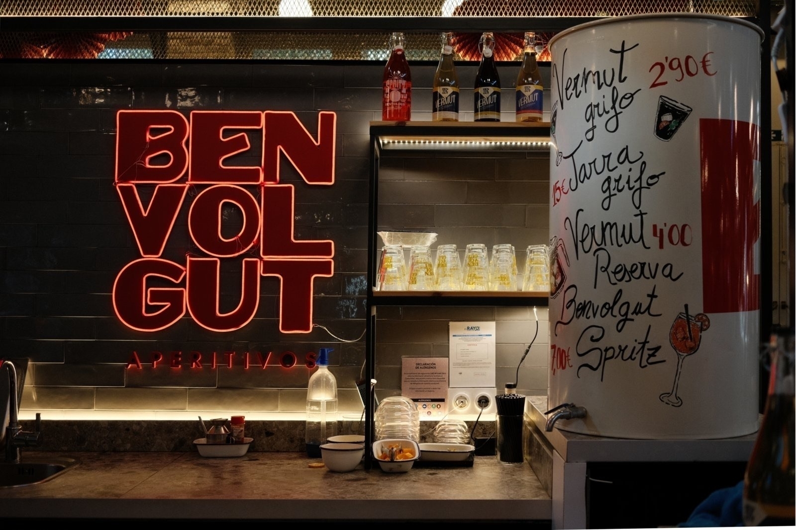 Auto-generated description: A bar counter features neon signage, drinks, and a menu showcasing aperitifs.