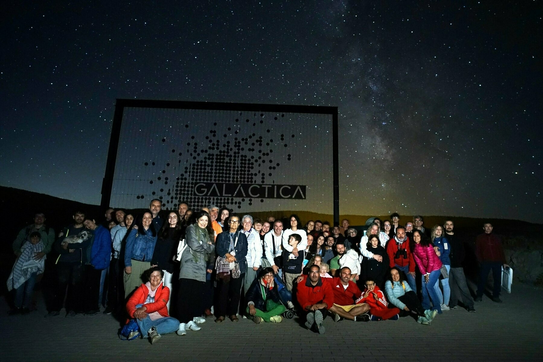 Auto-generated description: A group of people poses for a photo at night in front of a sign that reads GALÁCTICA, with a starry sky and the Milky Way visible in the background.