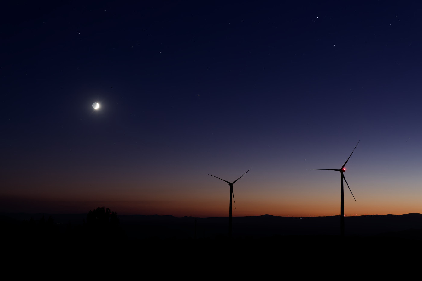 Auto-generated description: A serene dusk landscape features a crescent moon in a darkening sky with two silhouetted wind turbines.