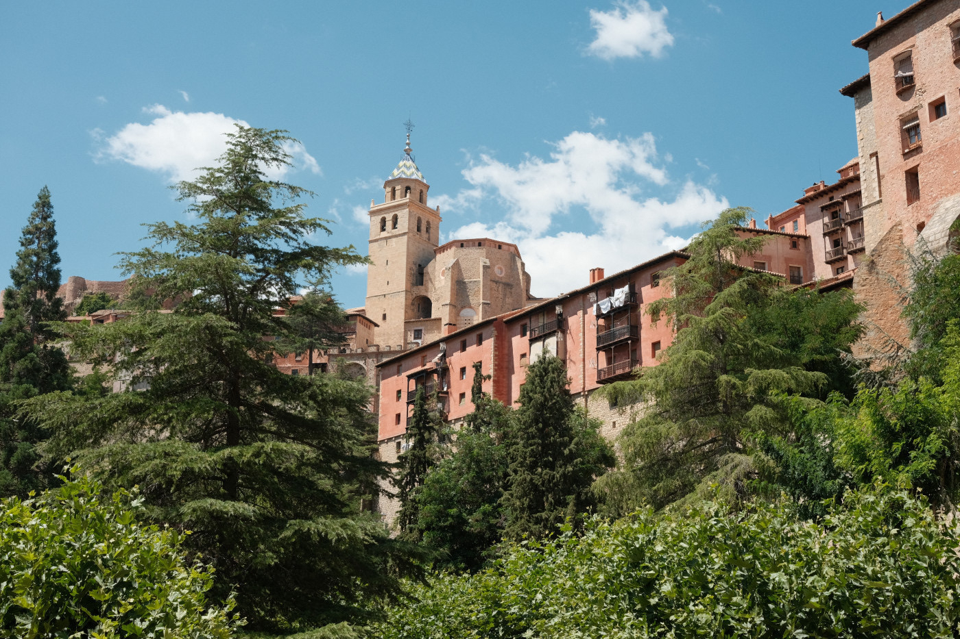 Auto-generated description: A picturesque village with red-roofed buildings and a prominent church tower is nestled among green trees under a blue sky.