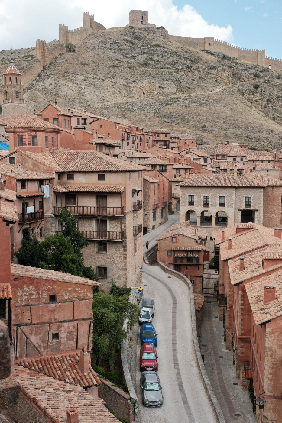 Auto-generated description: A picturesque town with terracotta buildings is nestled beneath a steep hill topped by a historic fortification wall.