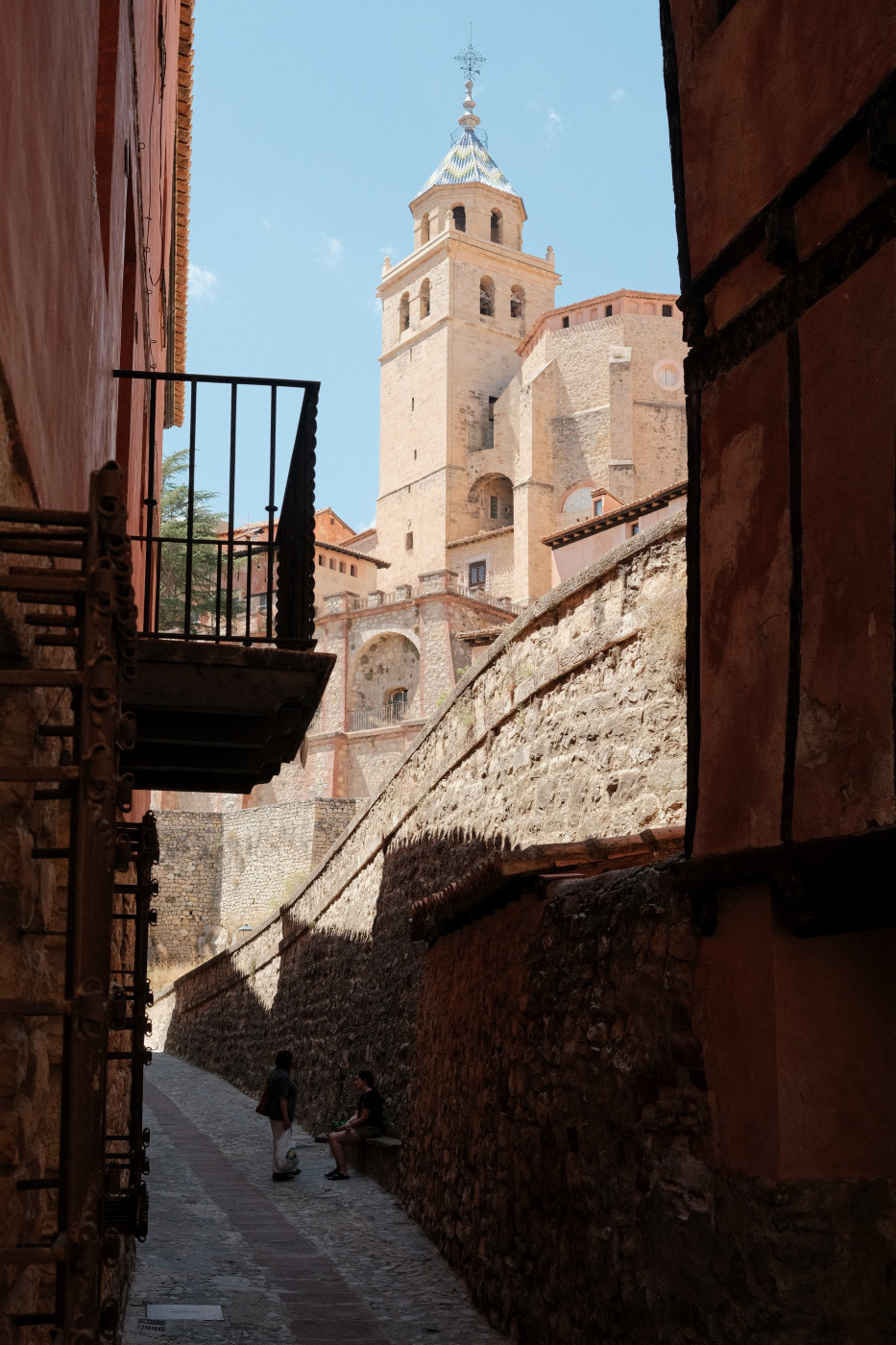 Auto-generated description: A narrow cobblestone street leads to a historic stone building with a tall tower, framed by shadowed walls and balconies.