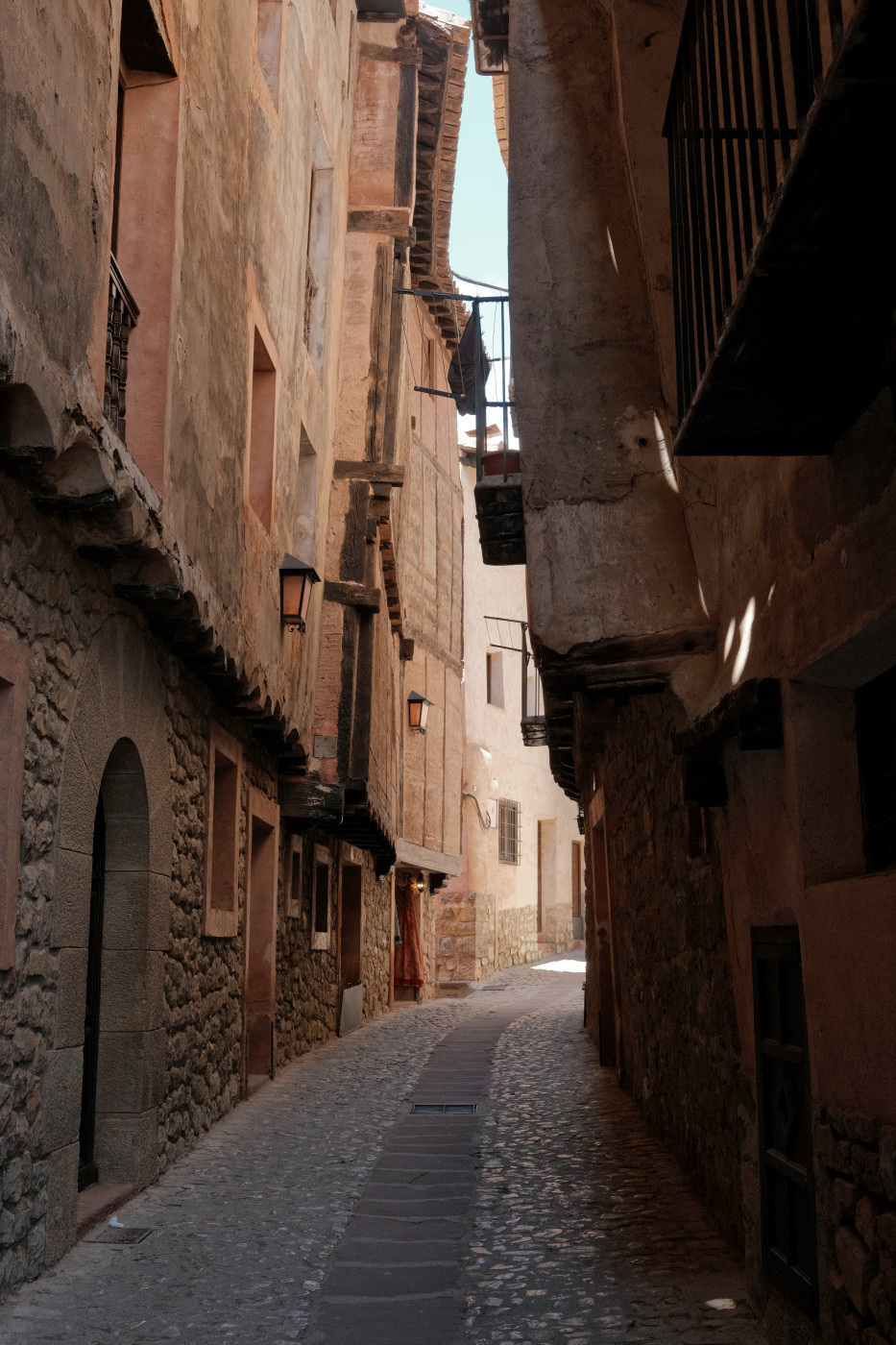 Auto-generated description: A narrow, cobblestone alleyway is flanked by tall, rustic buildings with wooden balconies and soft, warm lighting.