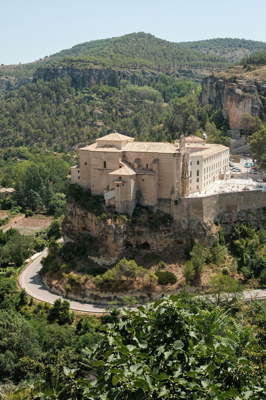 Auto-generated description: A large building with a beige facade is perched on a rocky hill surrounded by lush greenery, with a winding road encircling it.