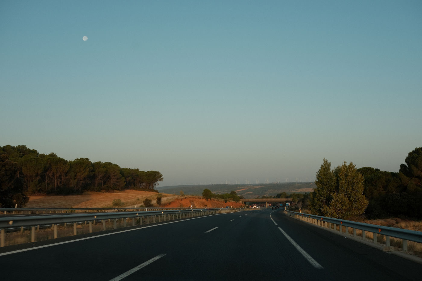 Auto-generated description: A highway stretches into the distance beneath a clear sky with the moon visible in the background, flanked by trees and rolling hills.