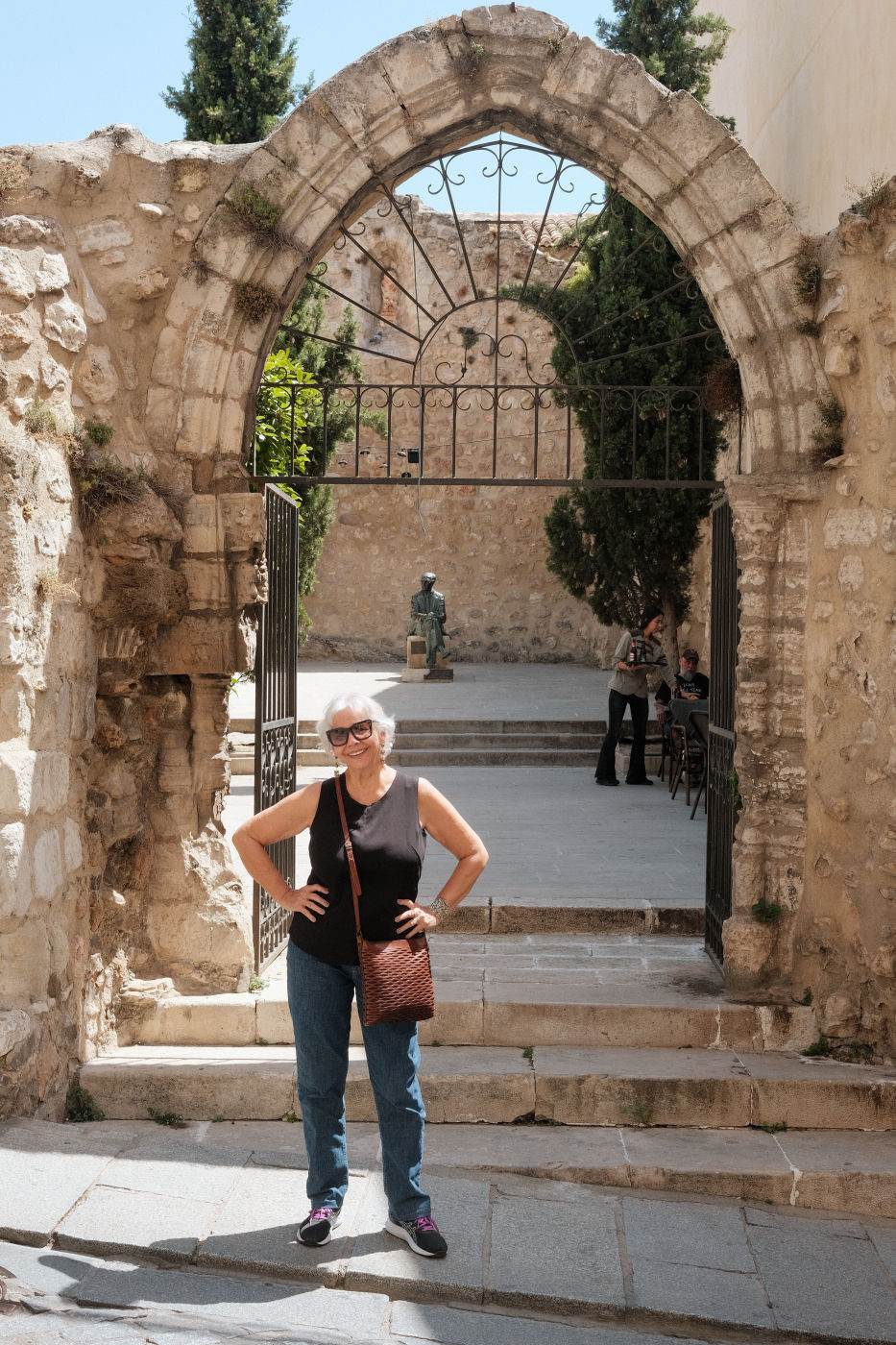 Auto-generated description: A person poses in front of an ancient stone archway with a metal gate, surrounded by trees and historical architecture.