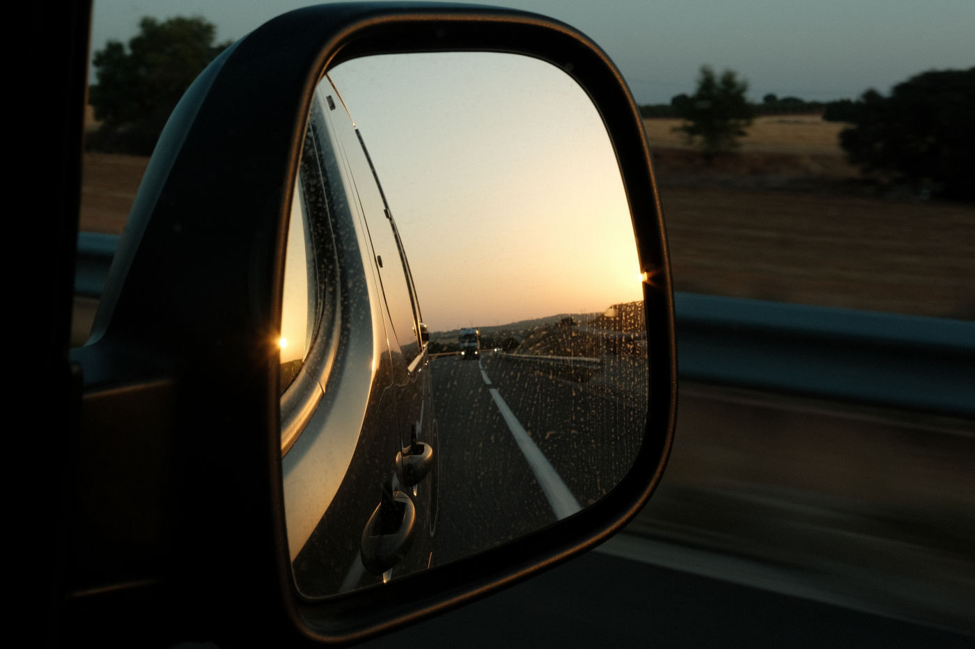 Auto-generated description: A rearview mirror reflects a sunset over a rural road with trees and fields in the background.