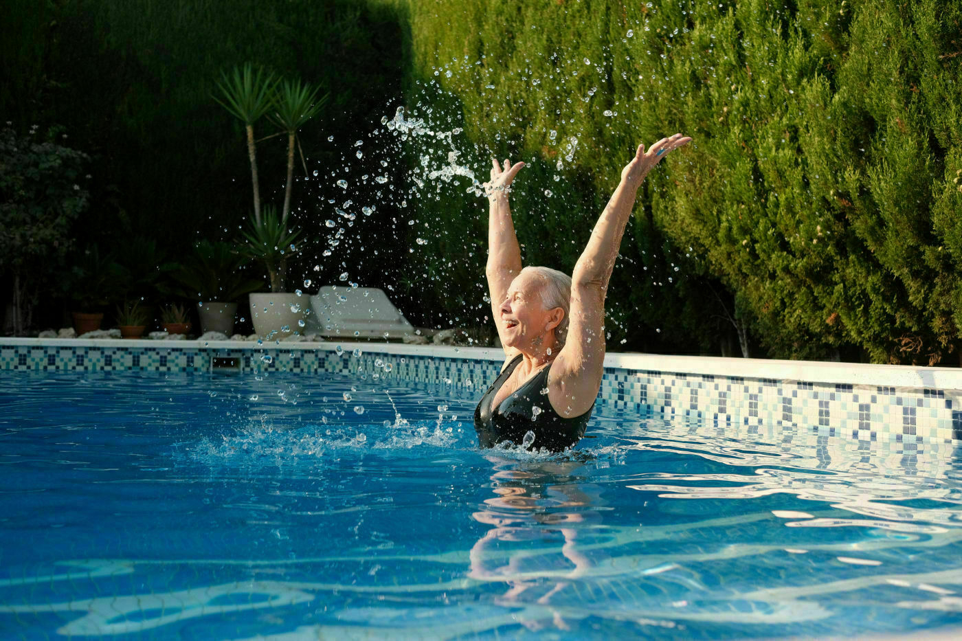 An older woman in a swimsuit joyfully raises her arms while splashing water in a swimming pool.