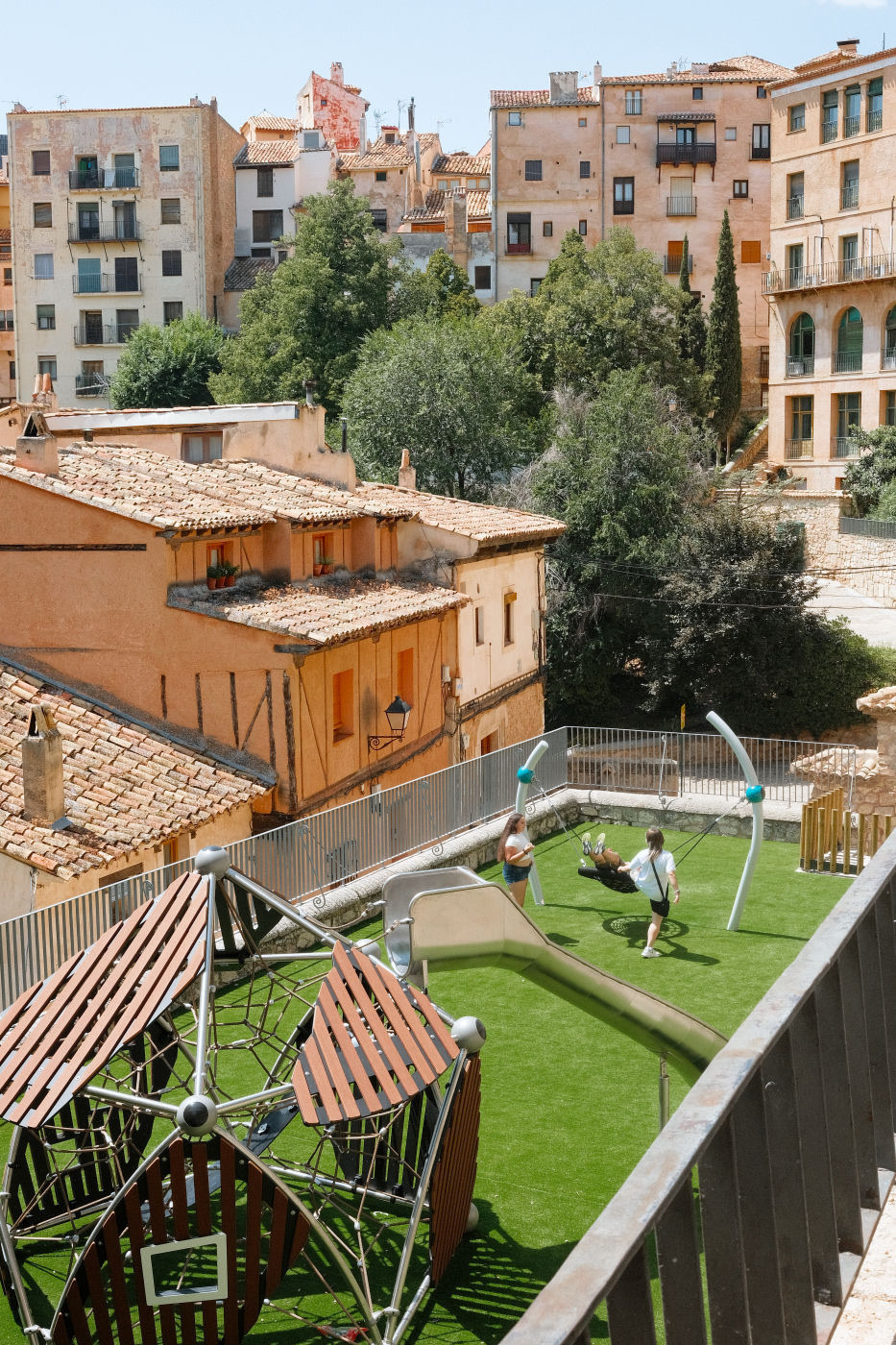 Auto-generated description: A playground with children on swings and climbing structures is set amidst old, terracotta-roofed buildings.